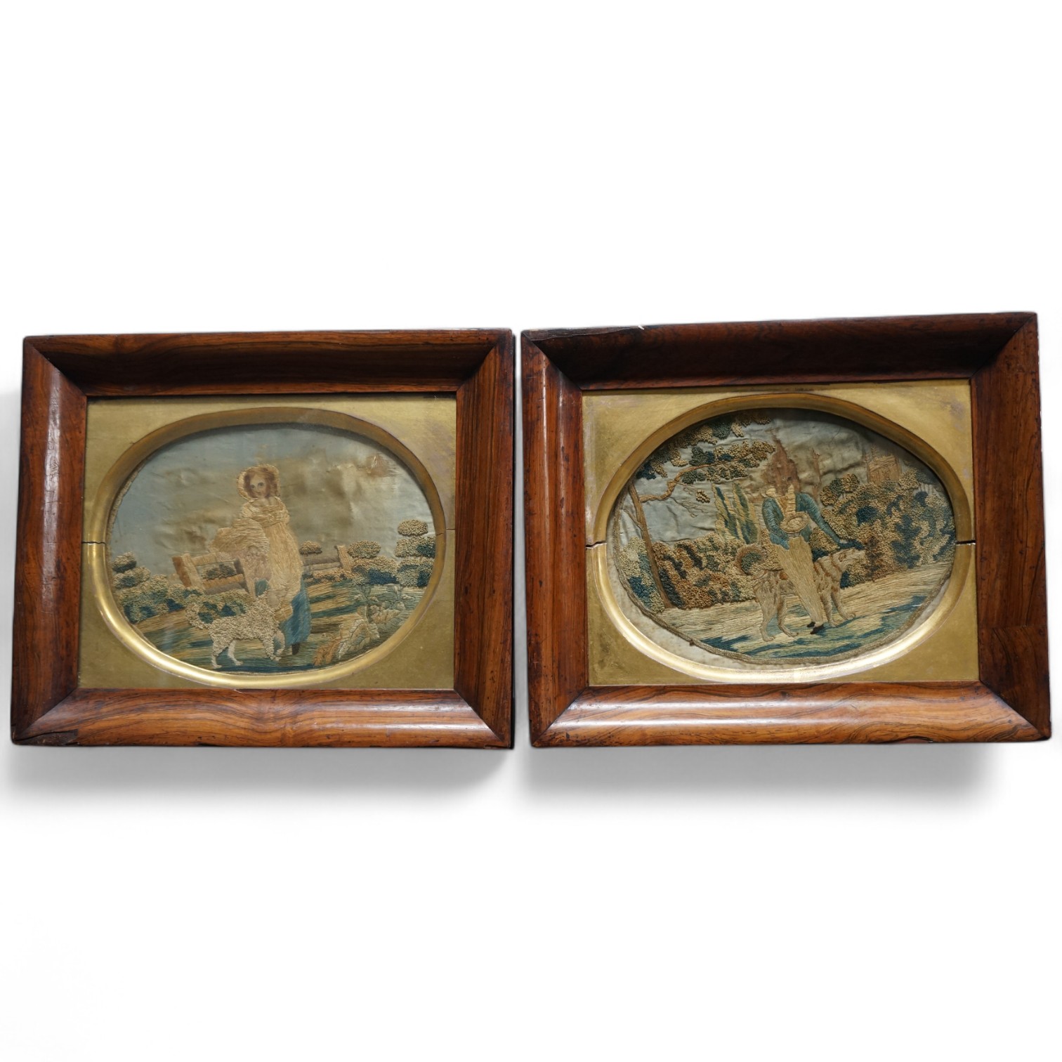 A pair of early 19th century rosewood-framed silk worked oval embroideries. Each embroidered with a boy and girl in a country landscape with their dogs. Worked in stem stitch and a knotting, the sky and faces hand painte